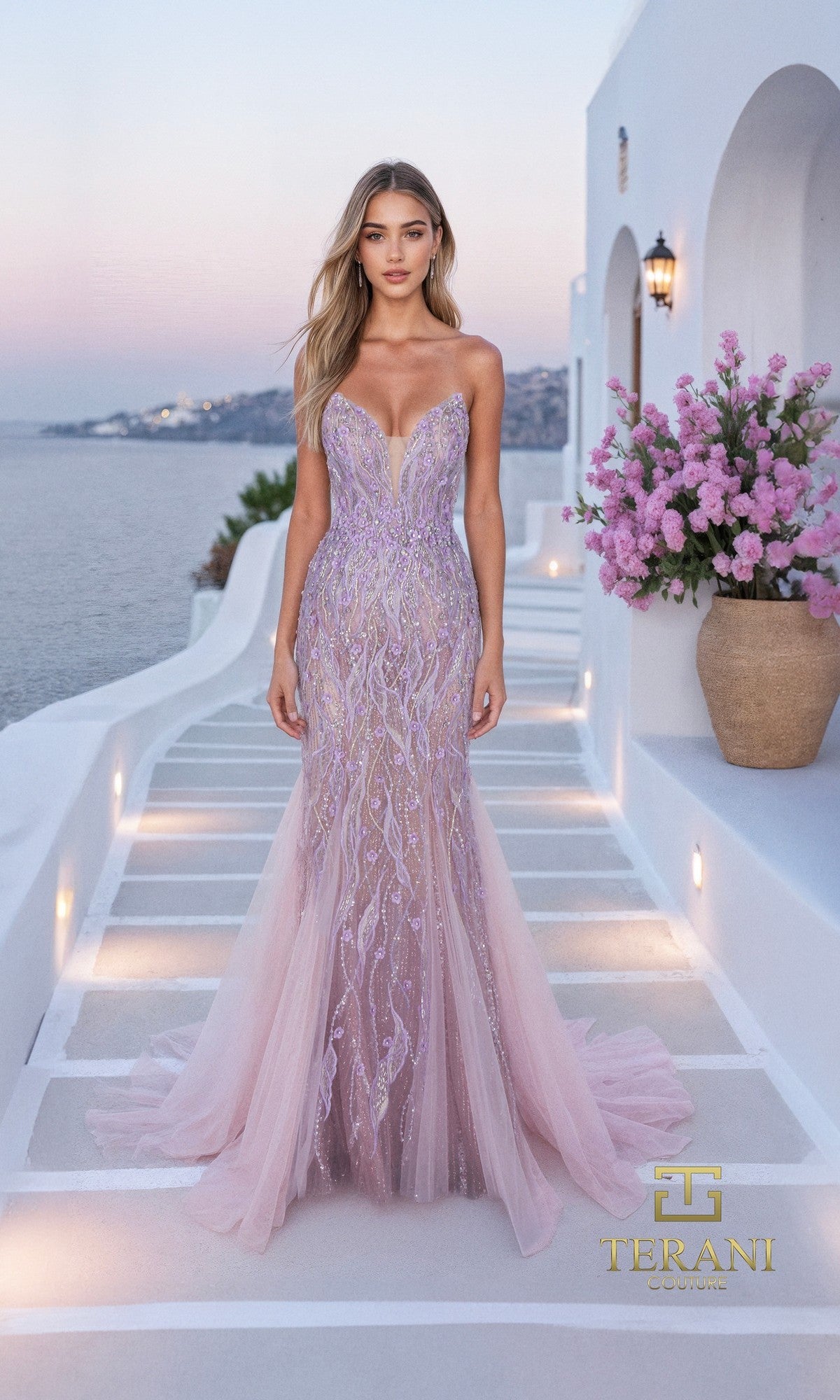 A woman in a fitted, strapless blush nude Long Formal Dress (Terani 261P6191) with embellishments stands on white stone steps by the sea, surrounded by flowers and Mediterranean-style architecture.
